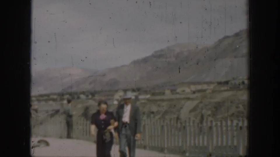 1937 California home-movie frame showing an adult couple on a paved dam overlook beside a white fence, with hills beyond.