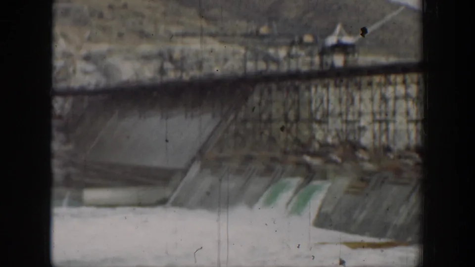 Faded 8mm frame of the California dam, showing railings, concrete spillway bays, and pale water beneath.