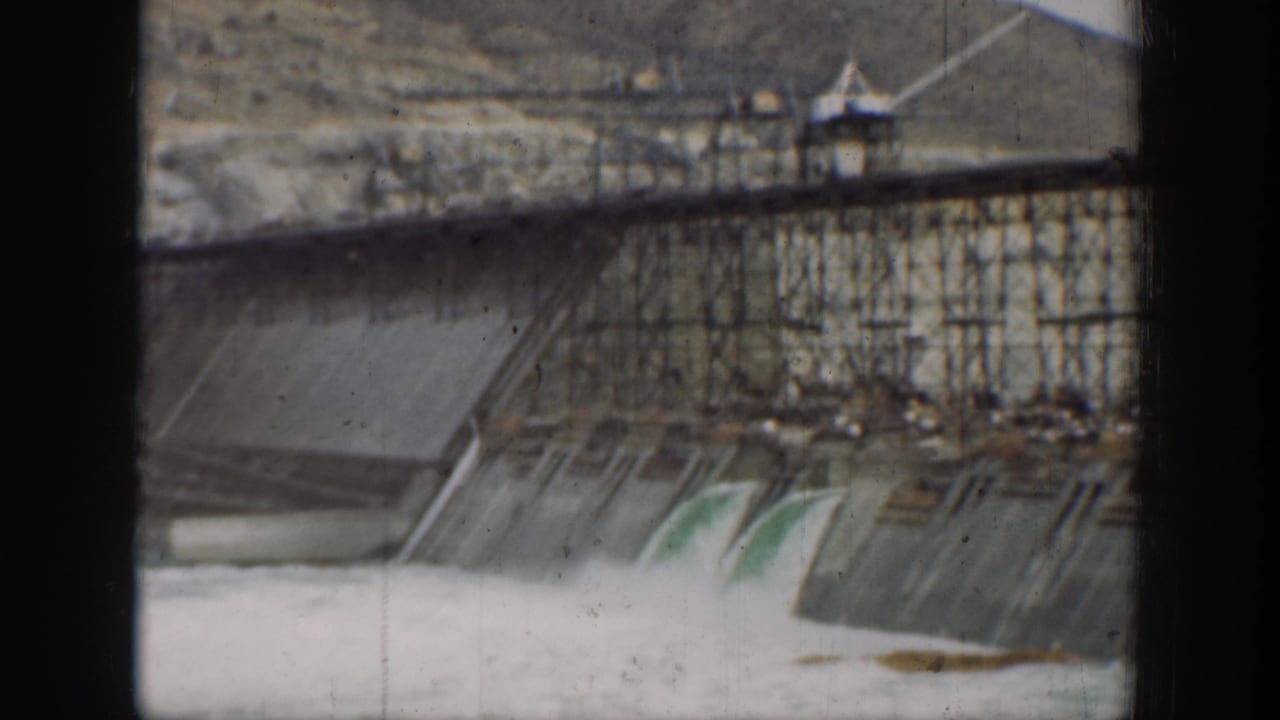 1937 California 8mm frame showing water pouring through a concrete dam spillway, with scaffolding and dark foreground bars framing the view.