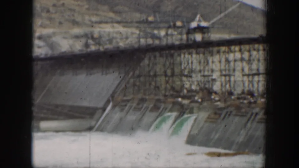 1937 California 8mm frame showing water pouring through a concrete dam spillway, with scaffolding and dark foreground bars framing the view.