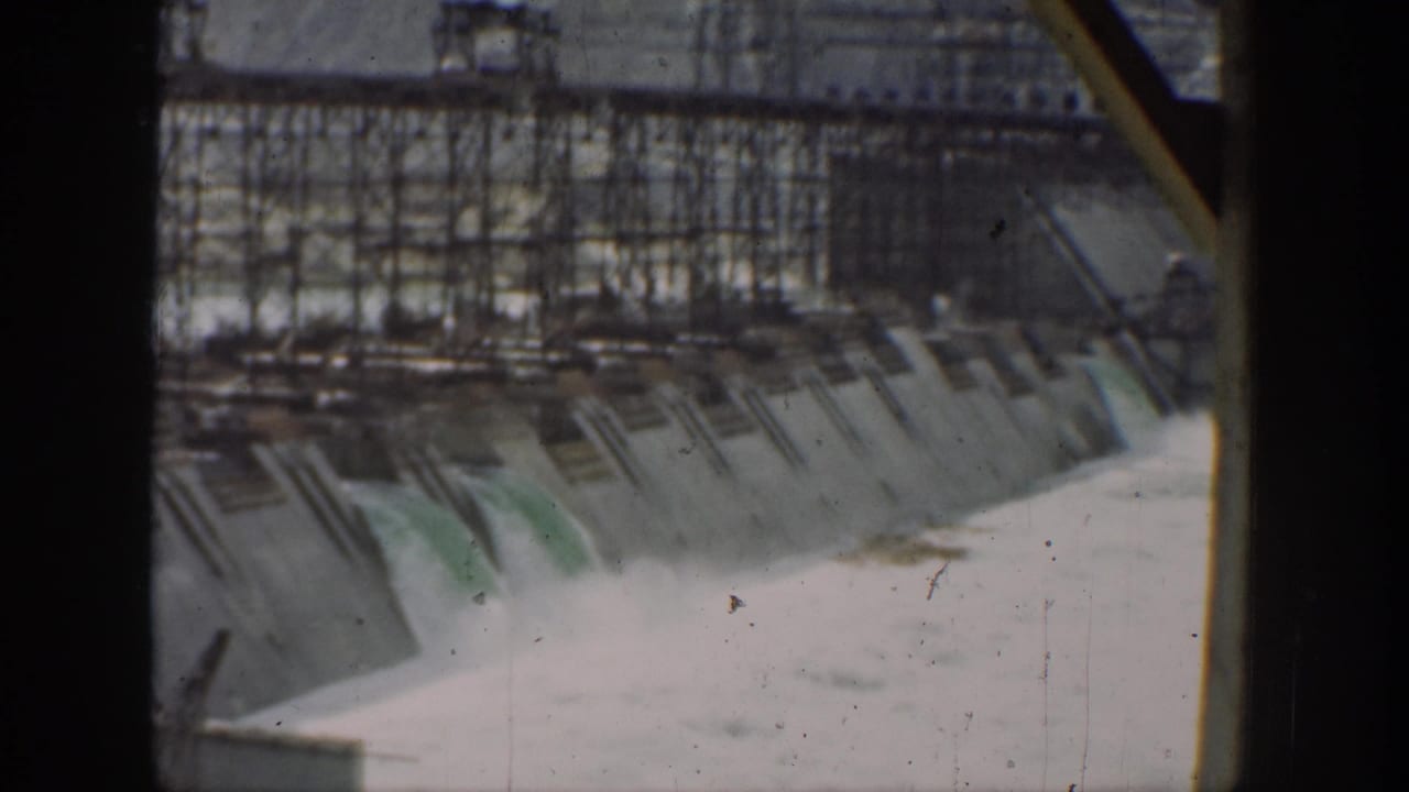 1937 California dam view with water flowing down spillway openings, scaffolding across the upper structure, and dark film-frame borders.