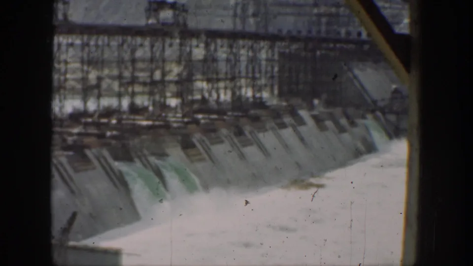 1937 California dam view with water flowing down spillway openings, scaffolding across the upper structure, and dark film-frame borders.
