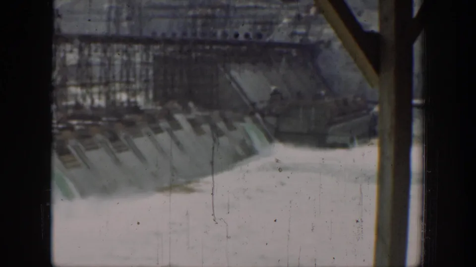 1937 California 8mm view of a dam spillway, showing pale water flow, concrete surfaces, scaffolding, and heavy dark foreground framing.