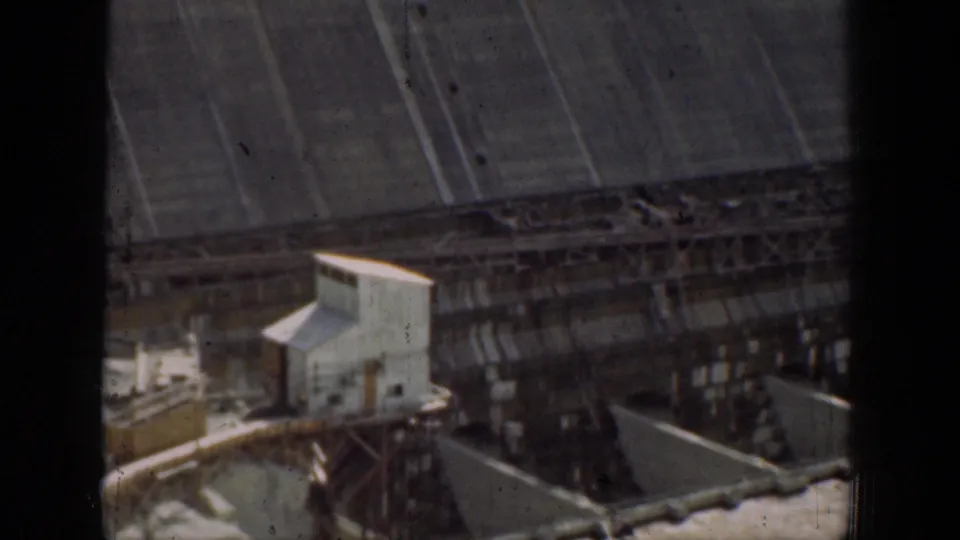 1937 California dam infrastructure frame showing small utility buildings, scaffolded concrete structures, spillway forms, and dark foreground framing.