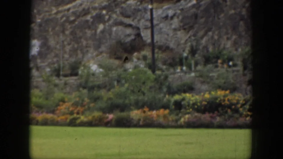 California 1937 home-movie frame showing a lawn and garden planting seen through dark window divisions, with rocky hills behind.