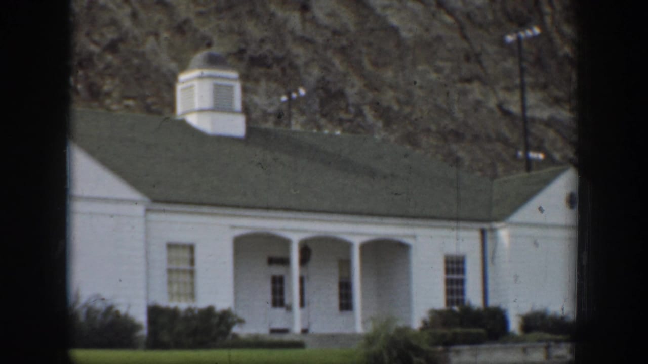 California 1937 frame of a small white house with a green roof and columned porch set against a rocky hillside.