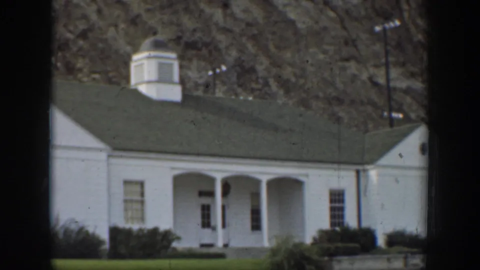 California 1937 frame of a small white house with a green roof and columned porch set against a rocky hillside.