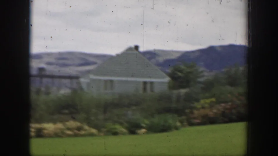 Soft 8mm view from California in 1937 showing a green-roofed house beyond flowers and lawn, with hills in the distance.