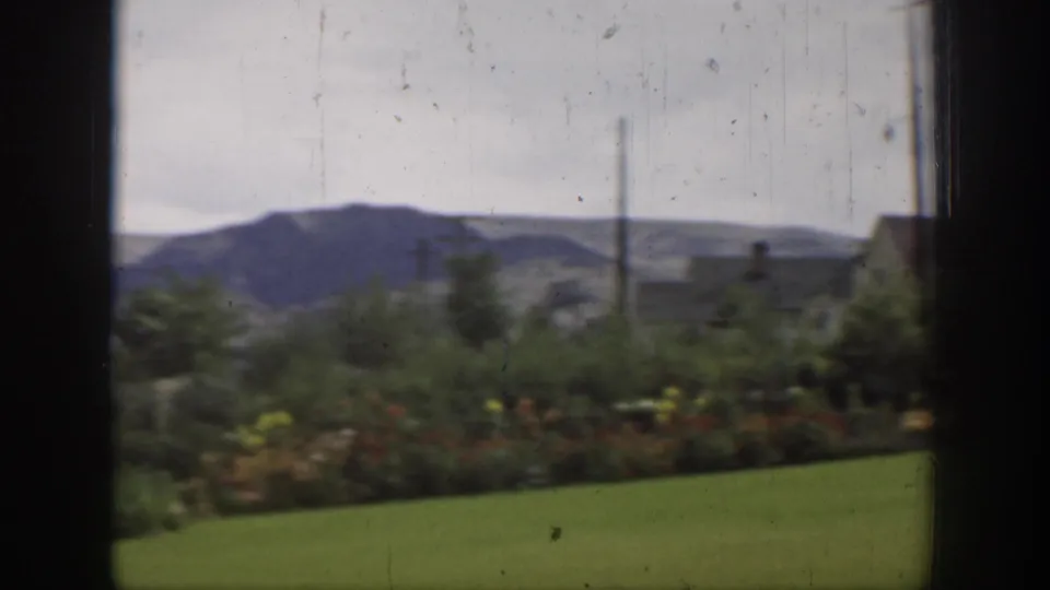 California 1937 home-movie frame of a sunny garden and lawn viewed through a window, with low mountains and poles beyond.