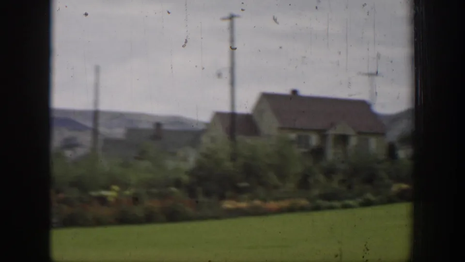 1937 California home-movie frame showing a rural house, lawn, shrubs, utility poles, and distant hills through dark vertical framing.