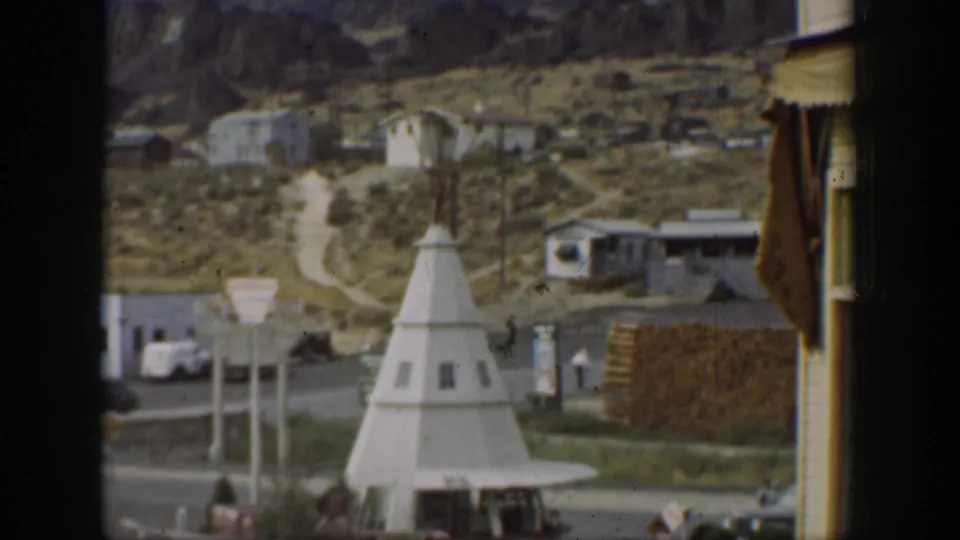 1937 California scene with a white teepee-shaped roadside structure, nearby signs, low buildings, parked vehicles, and hills in the background.