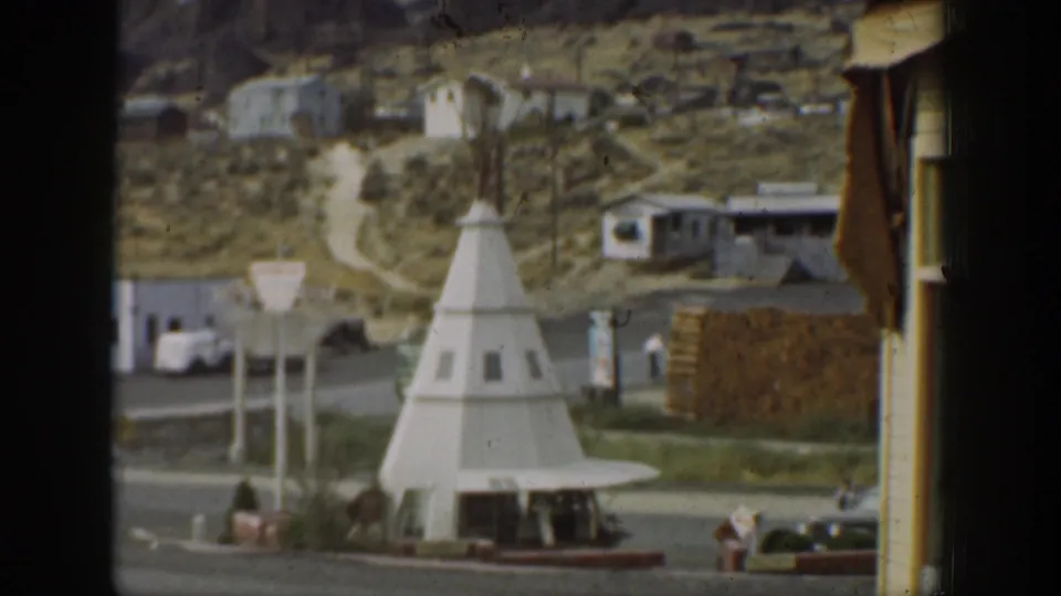 1937 California home-movie view of a white teepee-shaped building near a road, with hillside houses, signs, and parked cars around it.
