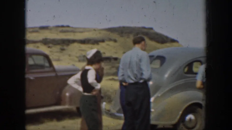 1937 California family outing frame with adults standing beside gray and brown 1930s cars against a sandy hillside backdrop.