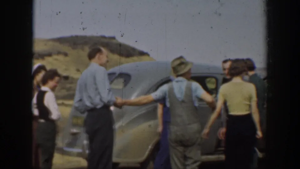 Adults in period clothing stand beside a silver sedan in rural California, with one handshake visible near the car.