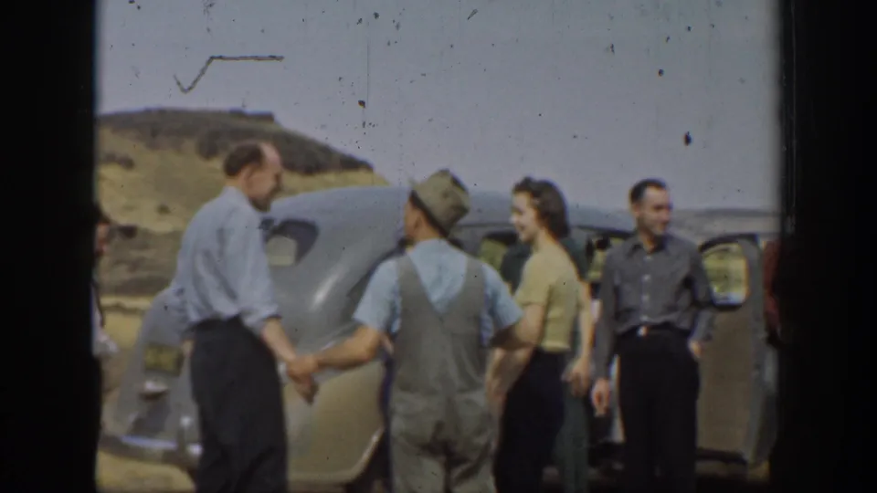 1937 California frame with adults gathered by a sedan, including a man in overalls and women in dresses near dry hills.