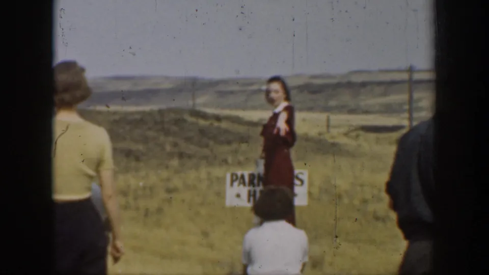 A woman in a dark dress stands near a small roadside sign as adults look across a dry California meadow landscape.