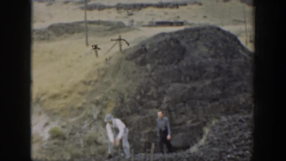 Two people in a dry California landscape near a large dark stone or coal-like pile, with a dirt track and low fencing behind them.