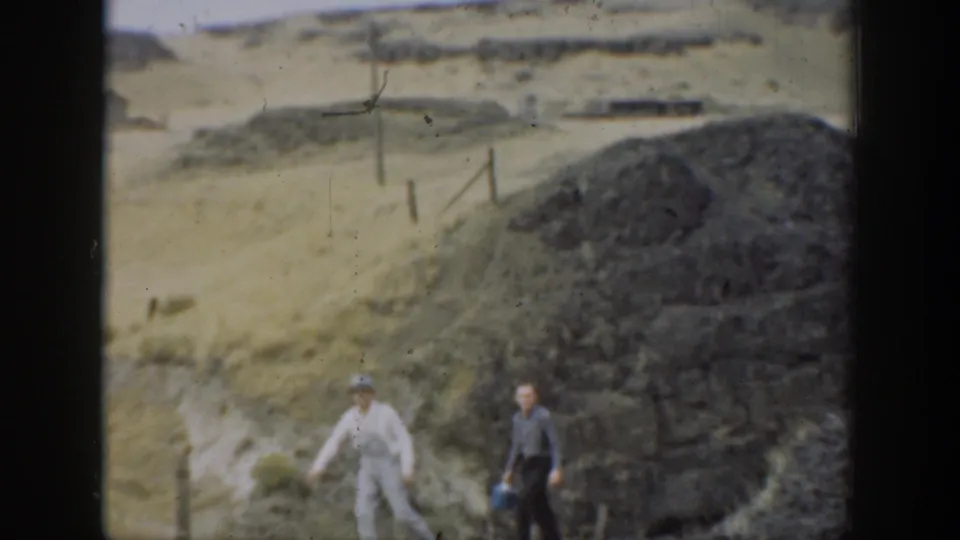 1937 California home-movie frame showing two people walking beside a dark stone pile, rough path, fence posts, and dry hills.