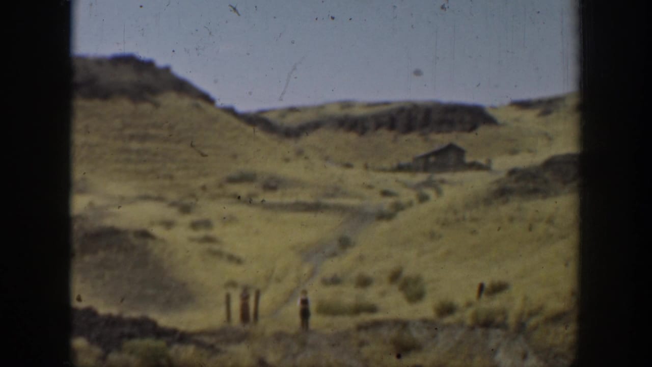 Dry hills with a small distant structure and a standing figure in open terrain.