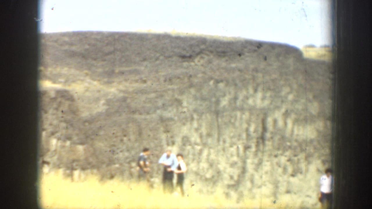 1937 California 8mm frame showing several visitors near a narrow waterfall at the base of a layered cliff under bright sky.