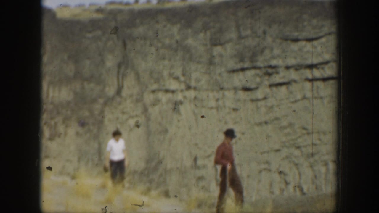 Two people stand apart in front of a tall, striated rock wall.