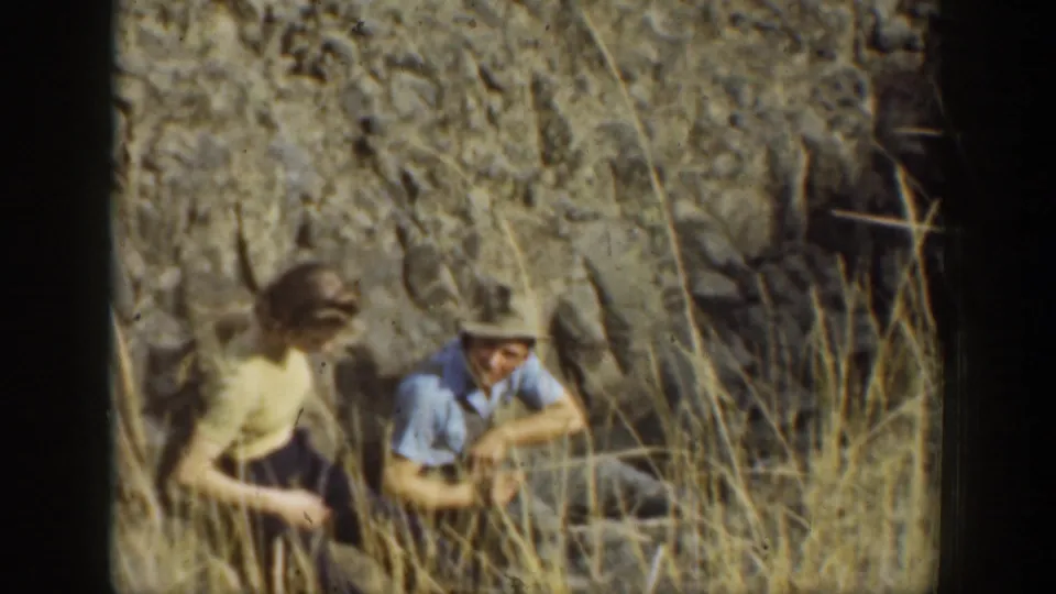 1937 California 8mm frame showing two visitors close to the ground among dry grasses beside a rugged rock wall.