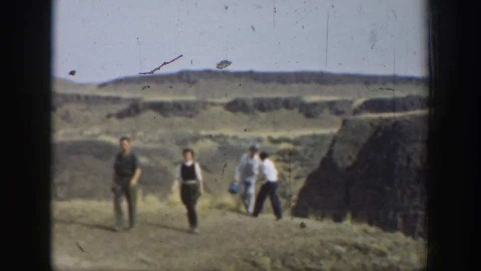 1937 California home-movie frame with several visitors walking through an open rocky area below distant layered hills.