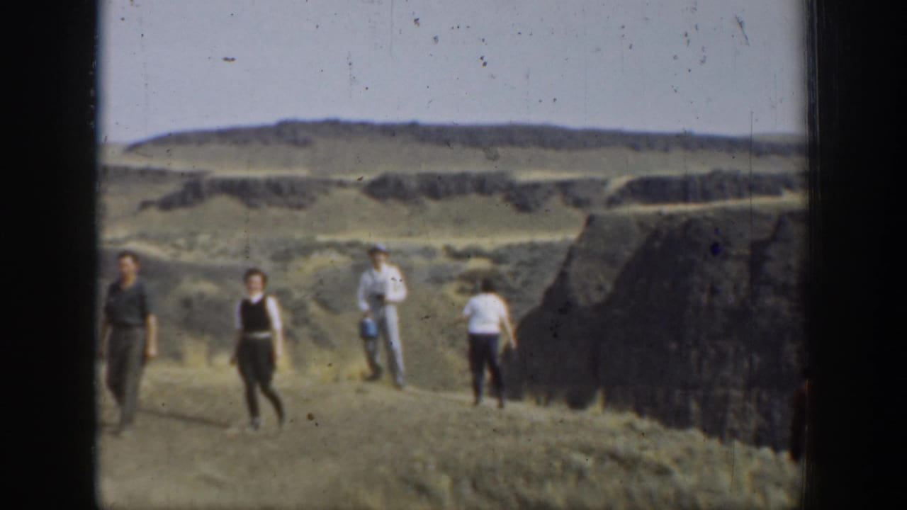 1937 California home-movie frame showing several people crossing uneven arid terrain near layered rock formations.