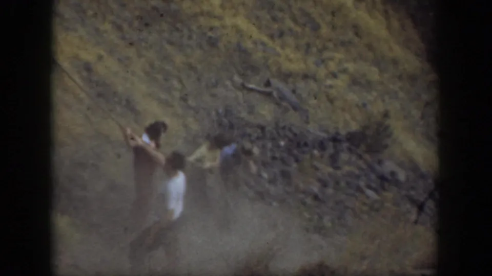 1937 California home-movie frame of a person in light clothing pulling or holding a rope near a dusty rocky dam face.