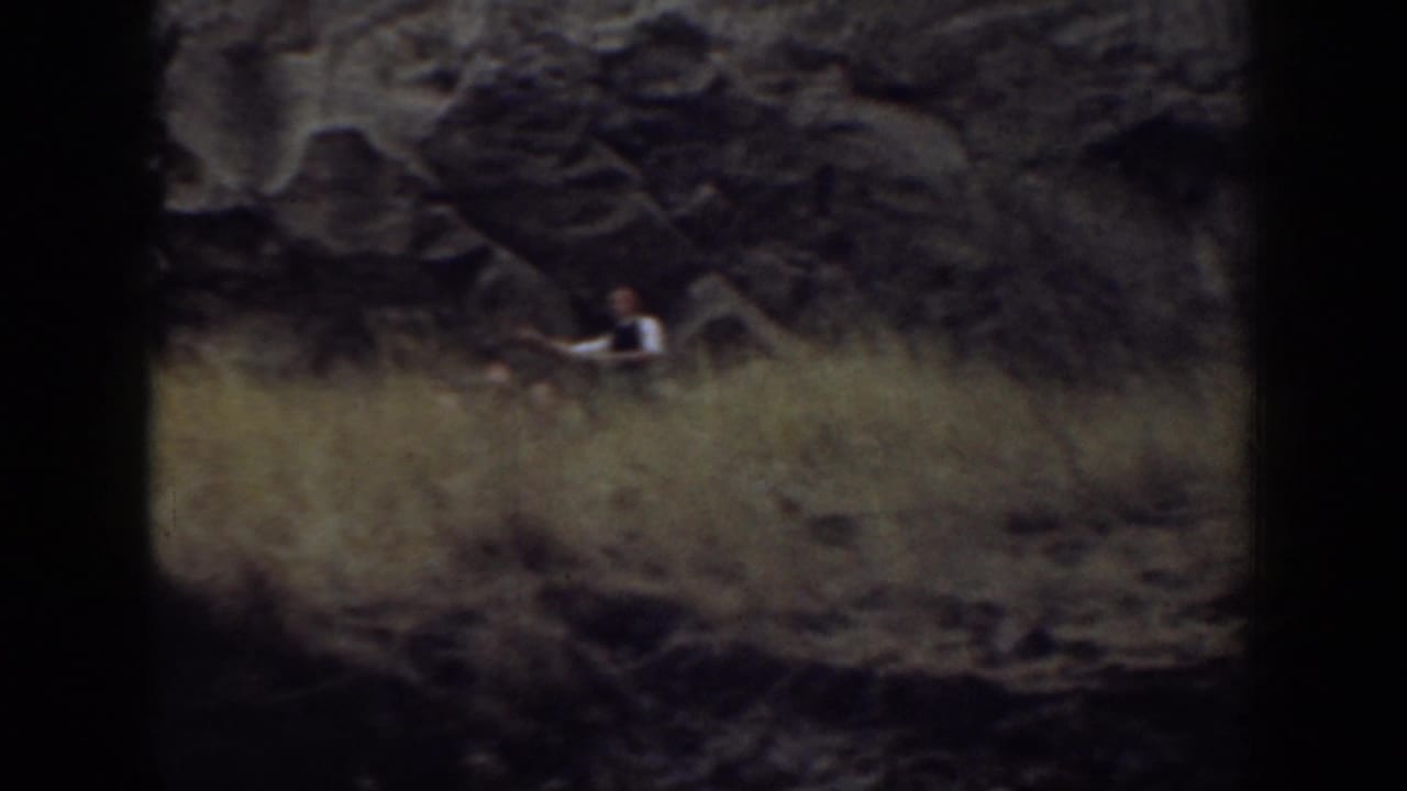 A distant adult sits or pauses near dark rock above tall dry grass.