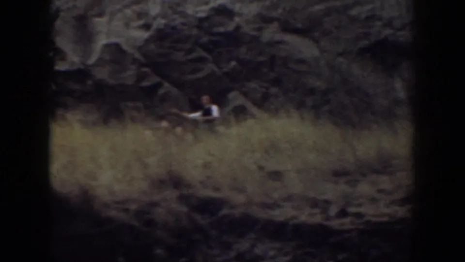 A distant figure pauses near a dark rock face above dry grass in the California hillside scene.