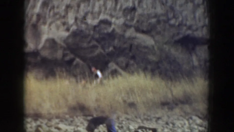 Adults are partly obscured by tall dry grass while moving below a rugged rock wall in the 1937 California footage.