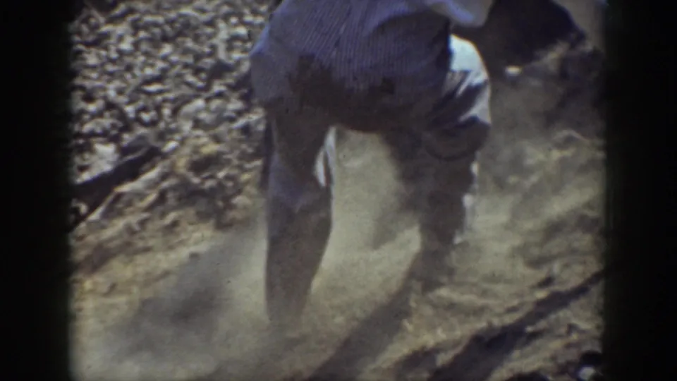 A close handheld frame shows a man climbing a dusty rocky slope, with loose stones and dry ground visible.