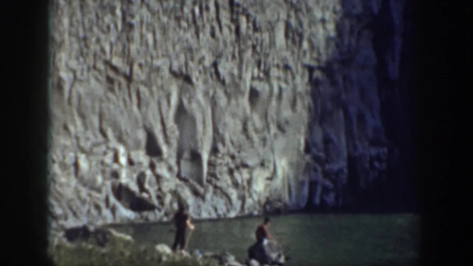Wide 8mm frame from 1937 California showing two people by a pool beneath a pale rock wall.