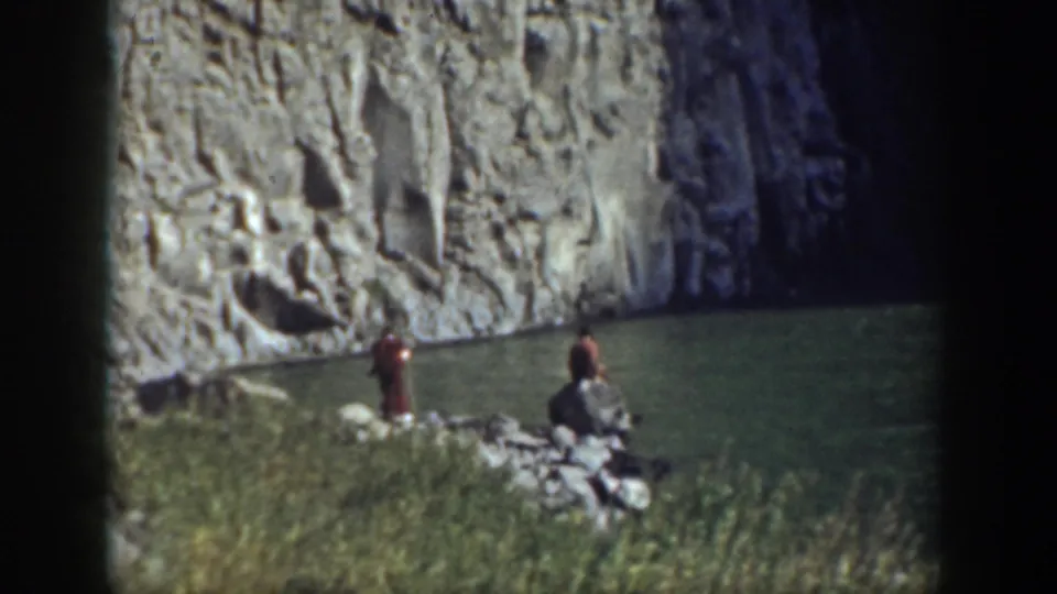 1937 California home-movie frame showing people near a quiet pool, with reeds in front and a rock wall behind.