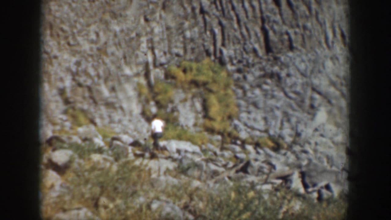1937 California home-movie frame showing a single visitor moving across rocks beneath a rugged cliff face.