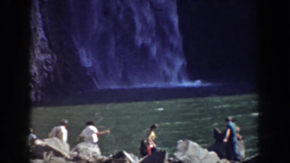 Tourists stand and sit on rocks near a green pool below a waterfall in 1937 California home-movie footage.