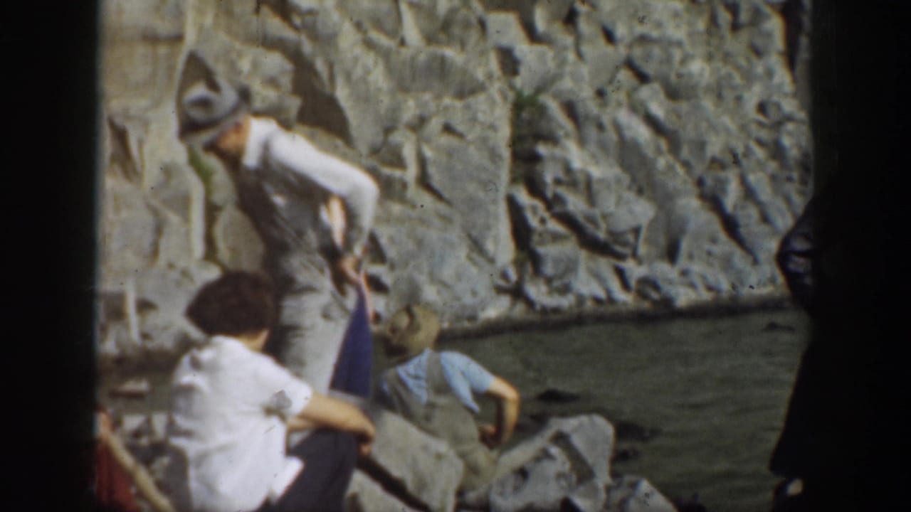 1937 California home-movie frame showing visitors on rocks near water, with a man in a light shirt and brimmed hat above seated figures.