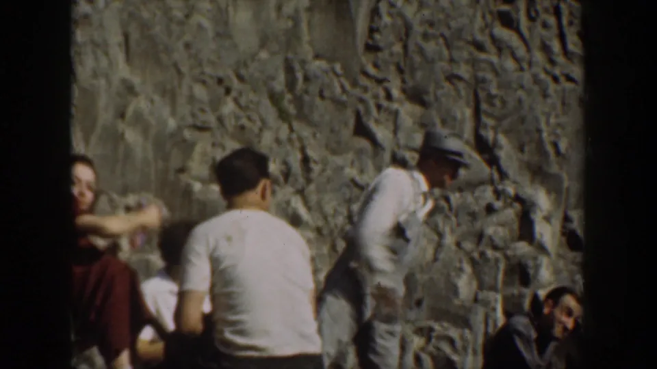 1930s California home-movie frame showing adults seated and standing on rocks near the waterfall area.