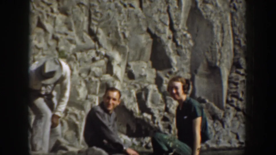 1937 California home-movie frame showing three adults resting beside a rugged rock wall in a mountain picnic setting.