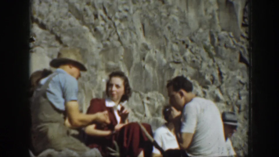 1937 California picnic frame showing seated adults by a rocky wall, including a hat-wearing figure and a woman in red clothing.