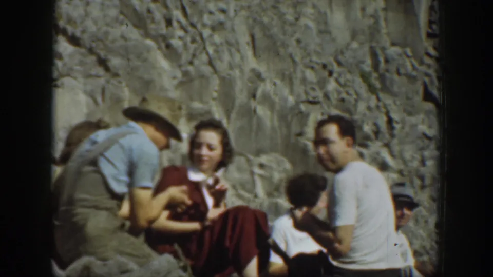 1937 California home-movie frame of a small picnic group seated beside a rocky cliff, with one man turned toward the camera.