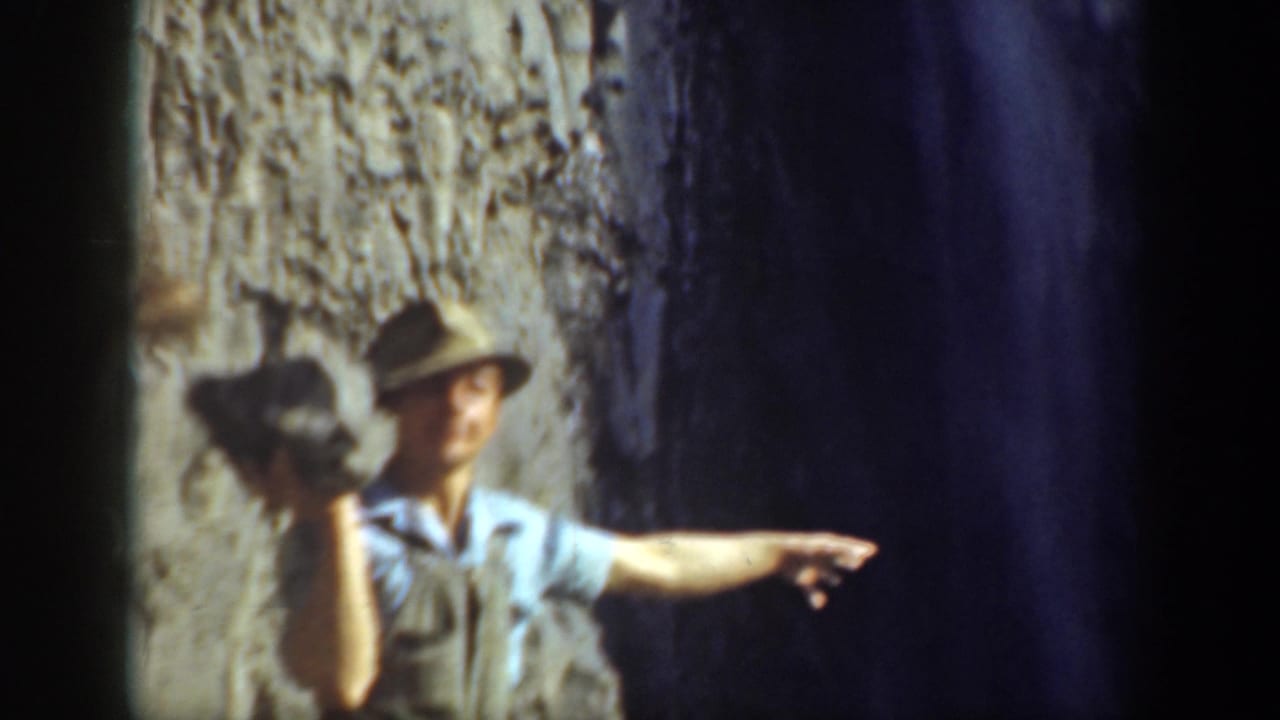 1937 California 8mm frame showing a man in a brimmed hat holding a rock and pointing near a rocky cliff and waterfall.