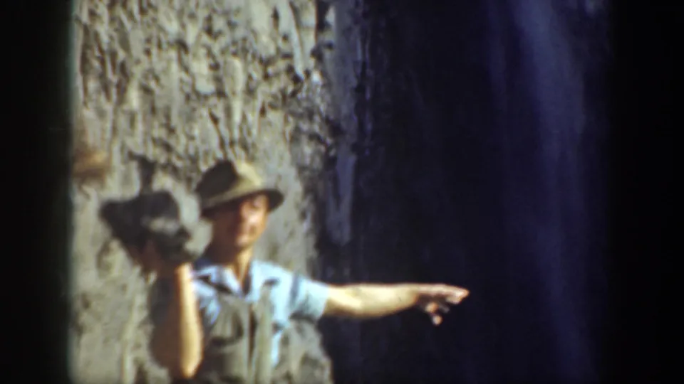 1937 California 8mm frame showing a man in a brimmed hat holding a rock and pointing near a rocky cliff and waterfall.
