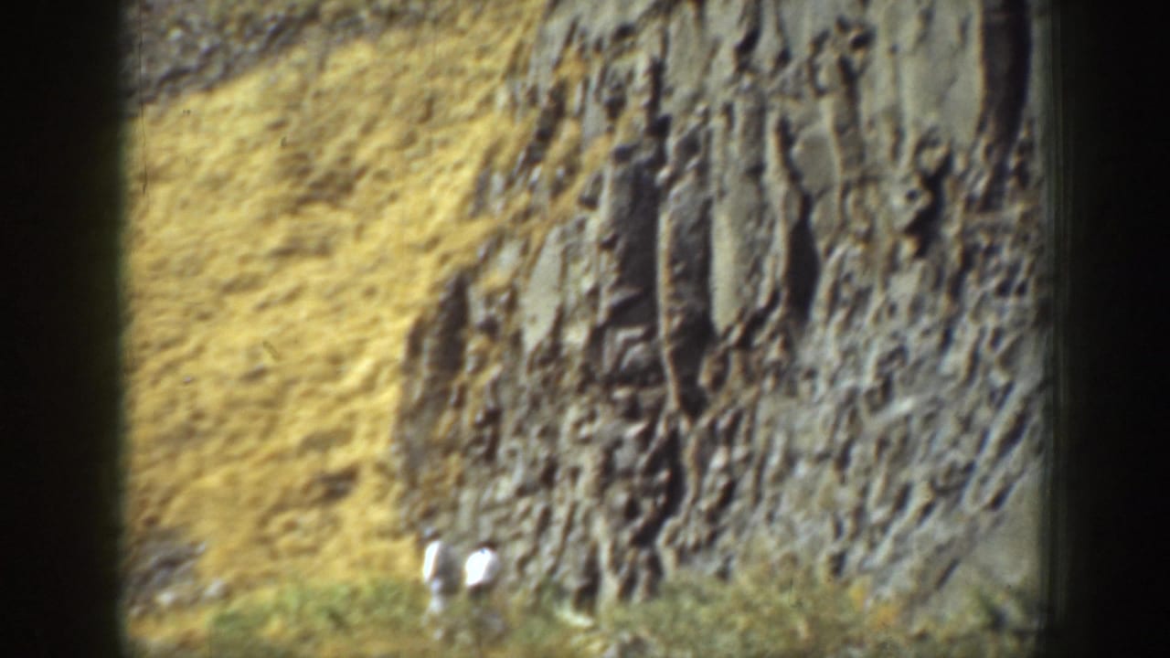 Two small figures stand near the base of a tall rock wall on a dry slope.