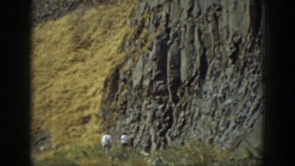 1937 California 8mm frame showing two distant hikers in light clothing near brush below a dark, jagged cliff face.