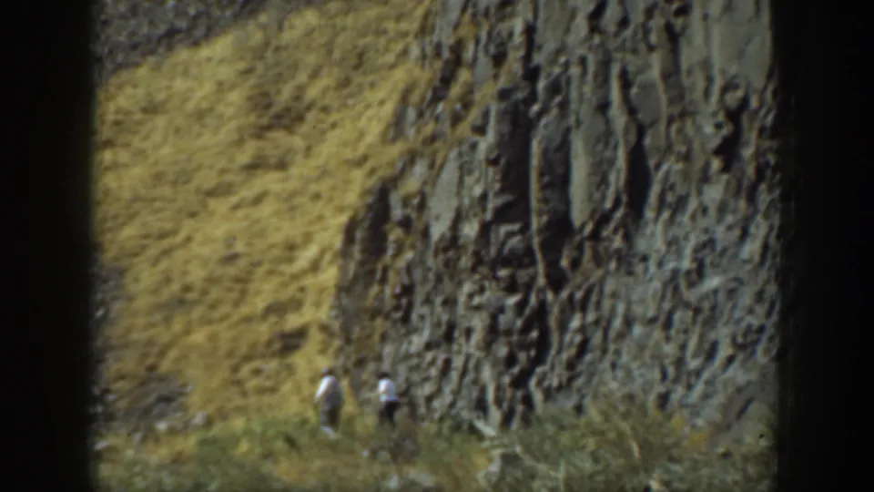 1937 California home-movie frame with two small hikers moving along a dry slope beside a textured rock formation.