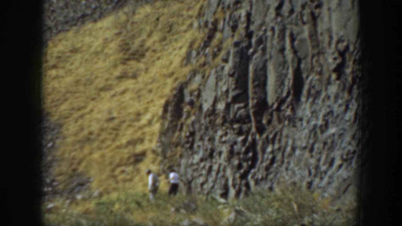 1937 California home-movie frame showing two adult men walking beside a steep, layered cliff with dry grass in the foreground.