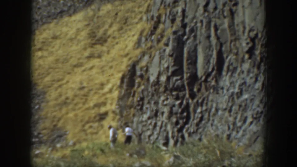 1937 California home-movie frame showing two adult men walking beside a steep, layered cliff with dry grass in the foreground.