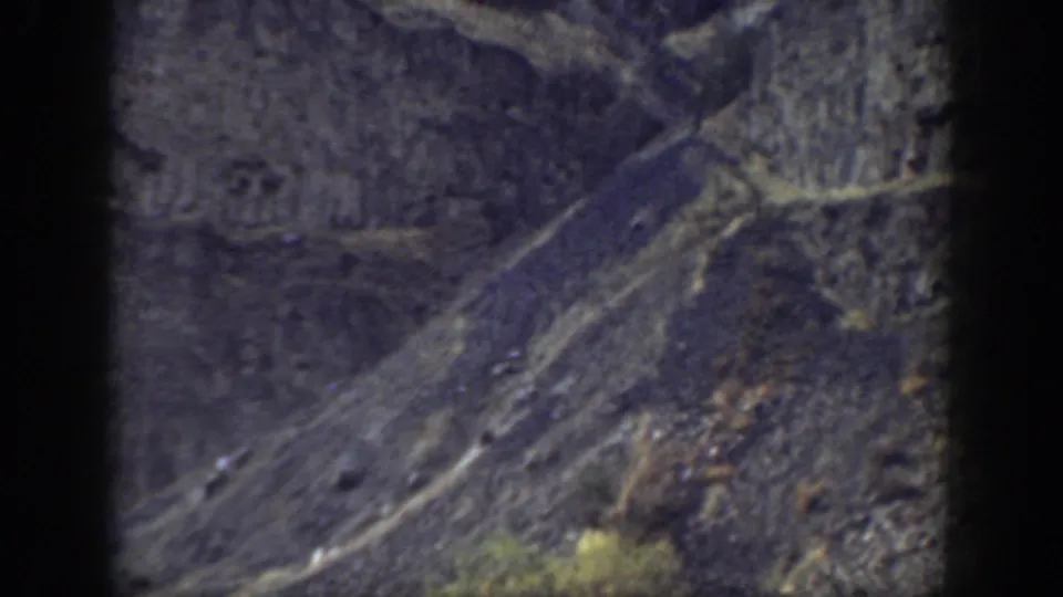 1937 California home-movie frame showing a steep mountain face with dark rock bands, scree, pale tracks, and scrub vegetation.